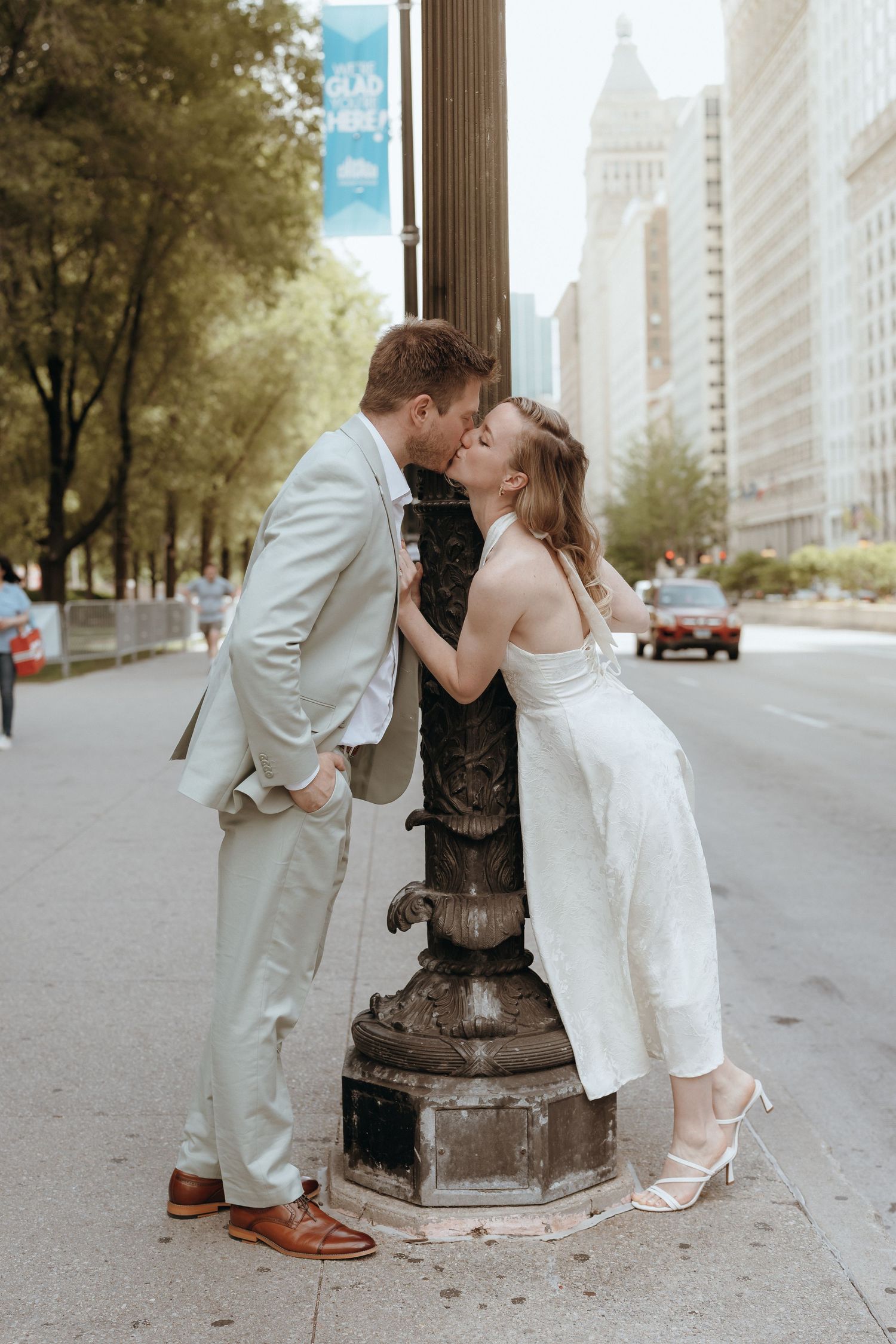 Bride and groom kissing beside an ornate street lamp on a city boulevard in wedding attire.