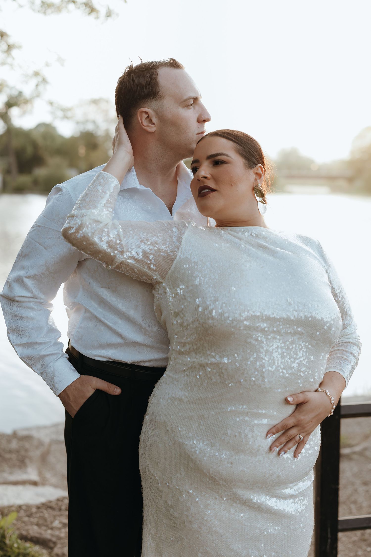 Groom in white shirt kissing bride in sequin wedding dress by a serene lakeside at sunset.
