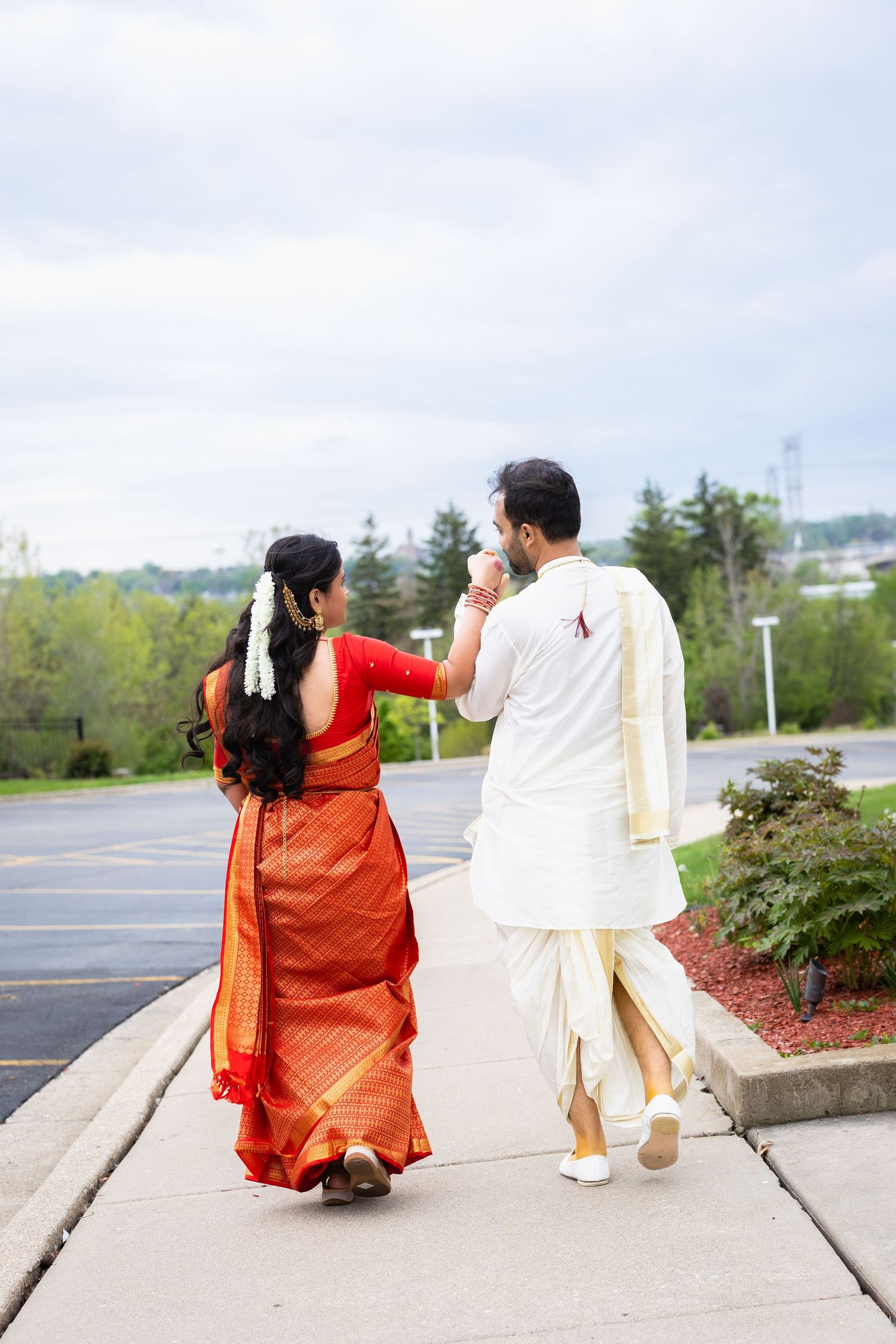 Indian couple in traditional attire walking hand-in-hand on a sidewalk, viewed from behind.