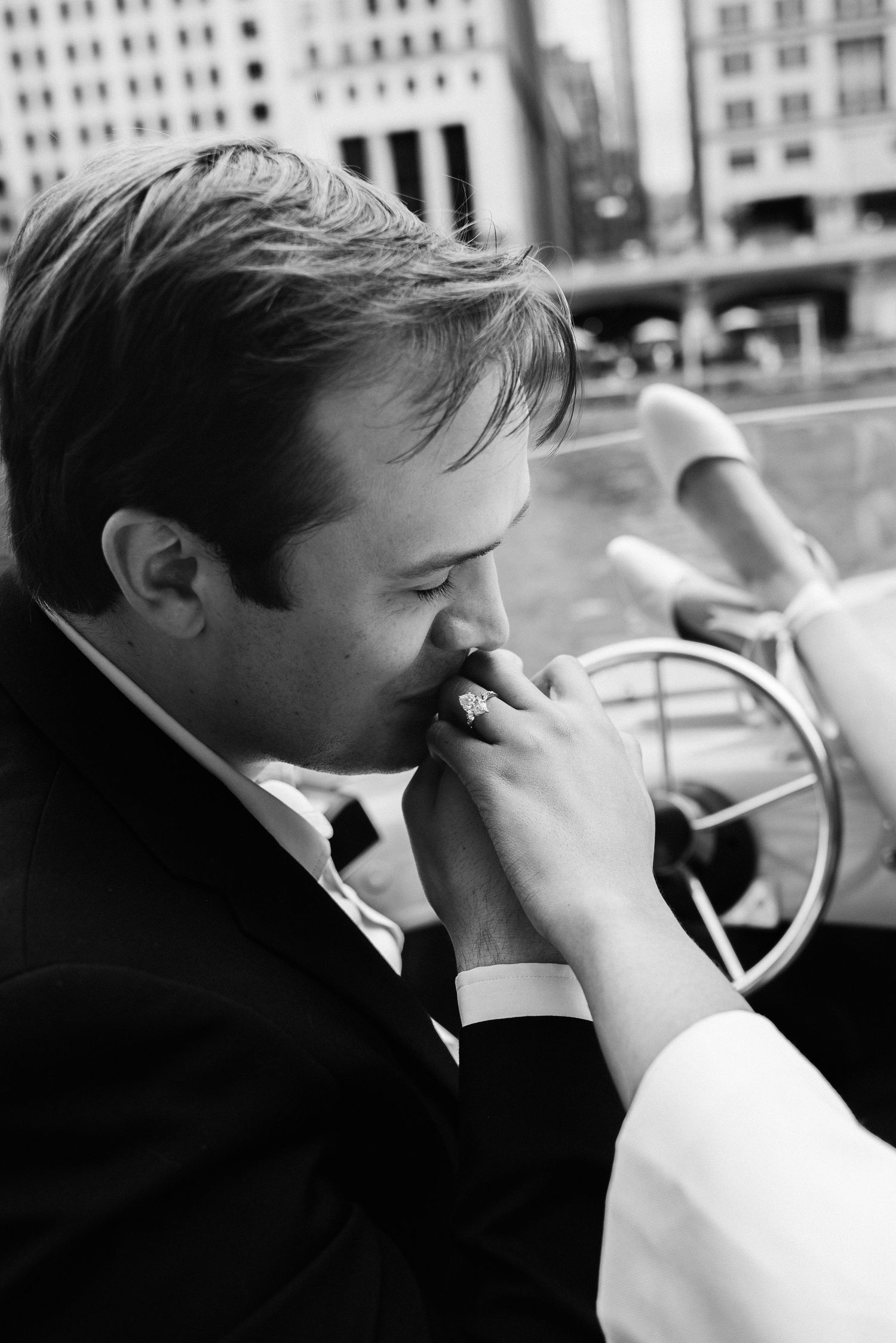 Groom in black suit kissing bride's hand with diamond ring in urban city setting, black and white photo.