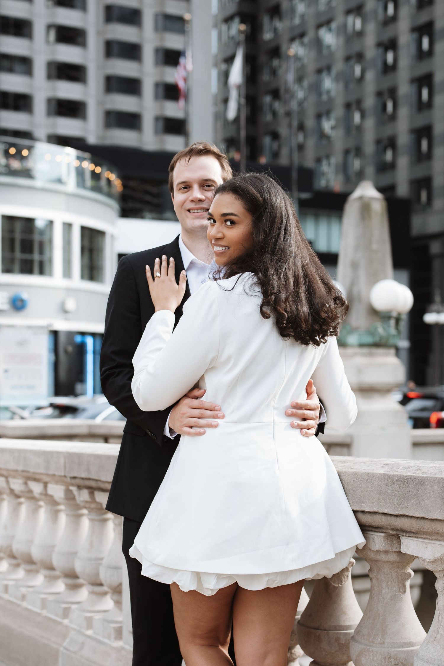 Happy interracial couple embracing on a city bridge, woman showing off engagement ring.