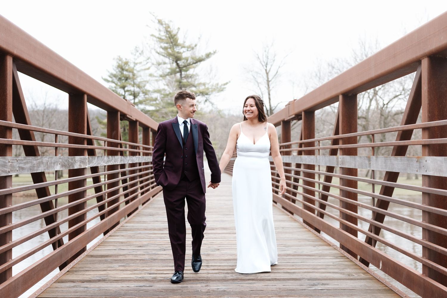 Bride and groom walk hand in hand on a wooden bridge, bride in white gown, groom in dark suit.