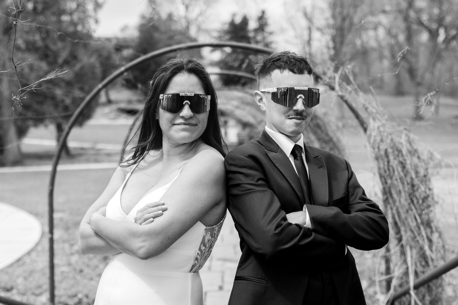 Black and white photo of a bride and groom posing back-to-back with arms crossed, wearing oversized sunglasses outdoors.