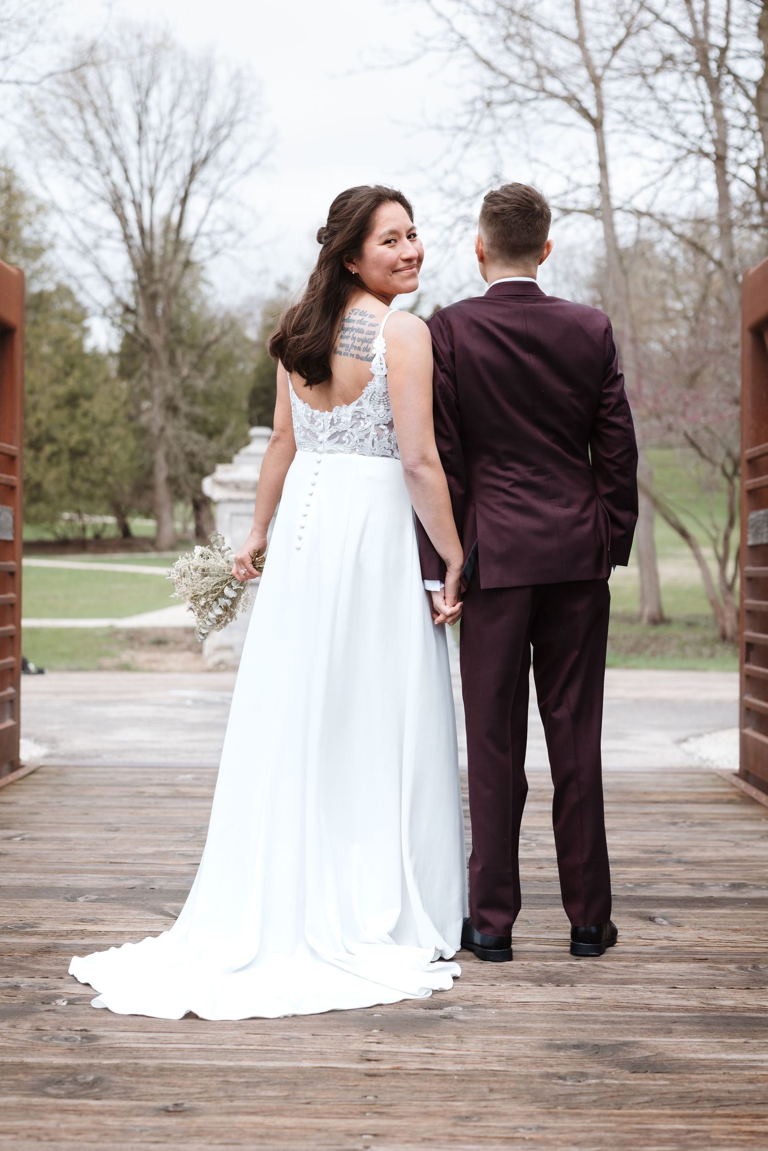 Bride in white gown holds bouquet, smiling back at camera while holding hands with groom in burgundy suit on wooden bridge.
