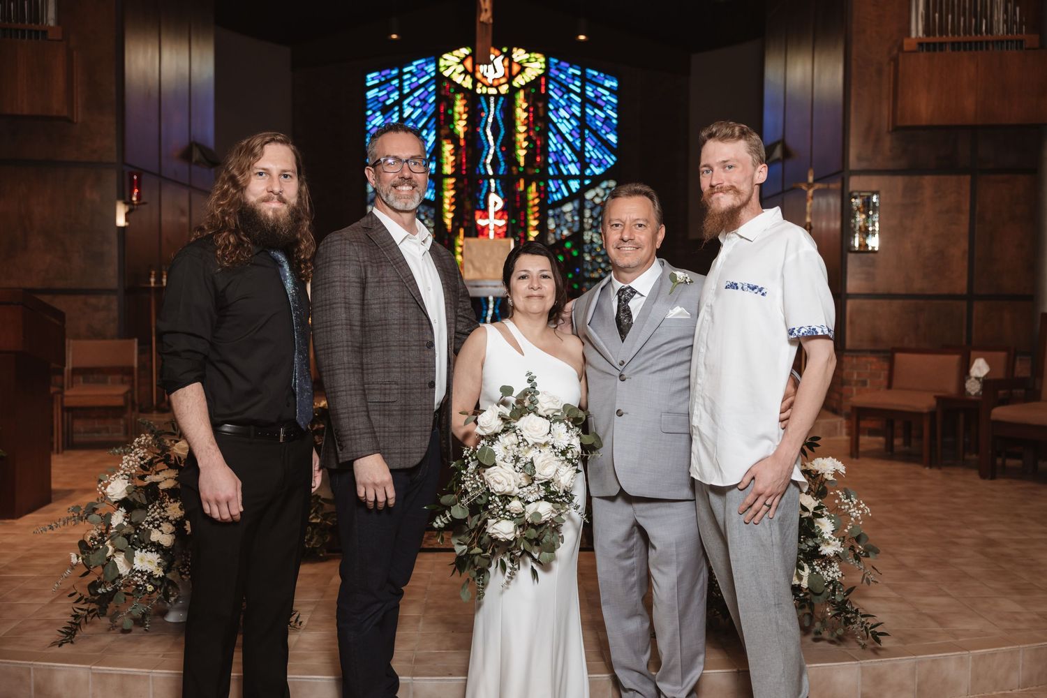 Wedding party of five poses together in front of colorful stained glass windows inside a church.