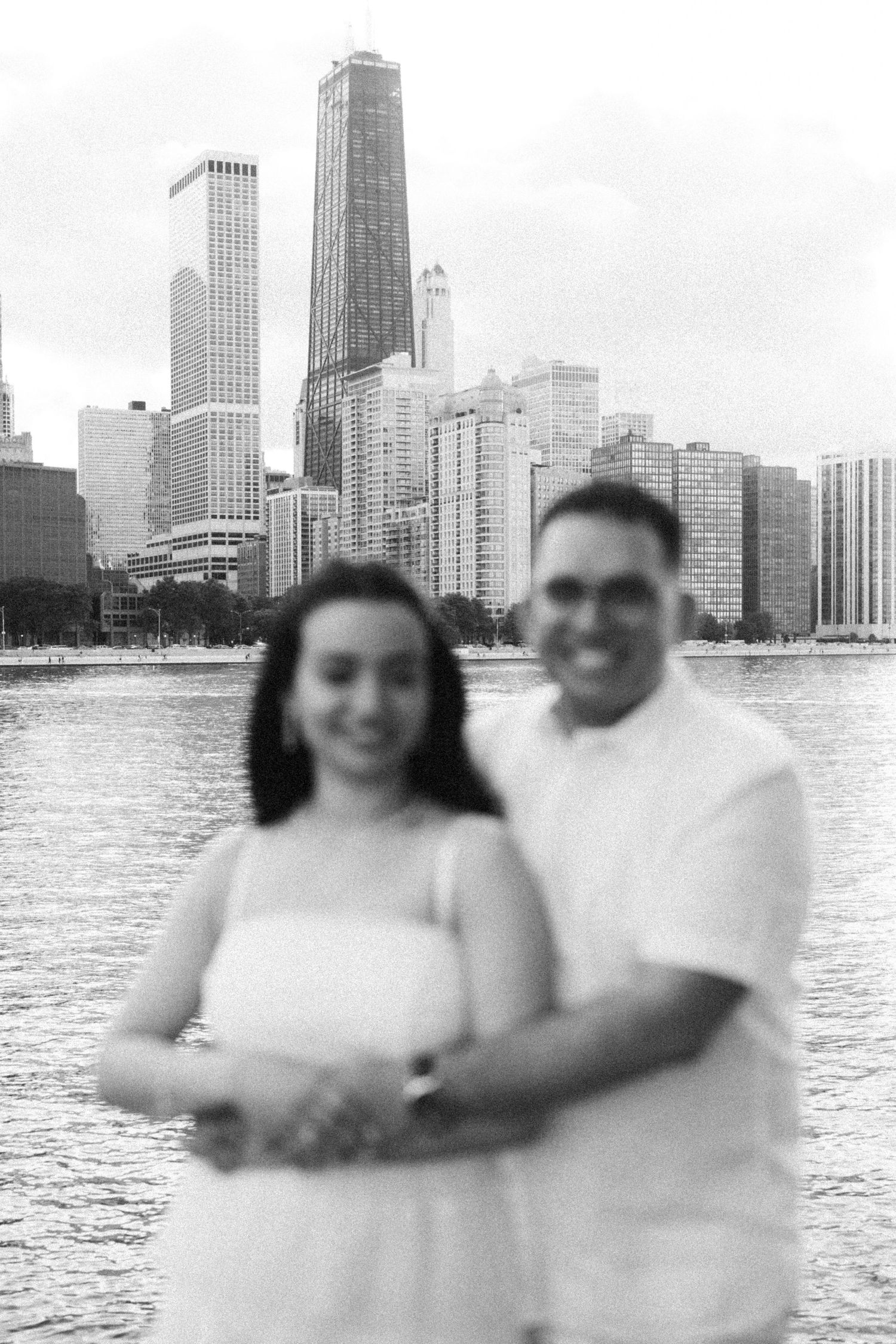 Black and white photo of a couple posing in front of the Chicago skyline across the water, slightly out of focus.