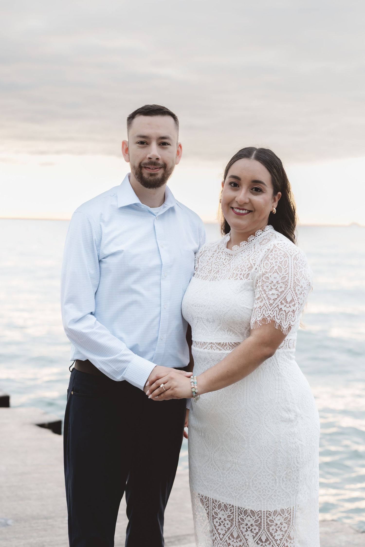 Couple poses romantically by the ocean at sunset, woman in white lace dress, man in light blue shirt.