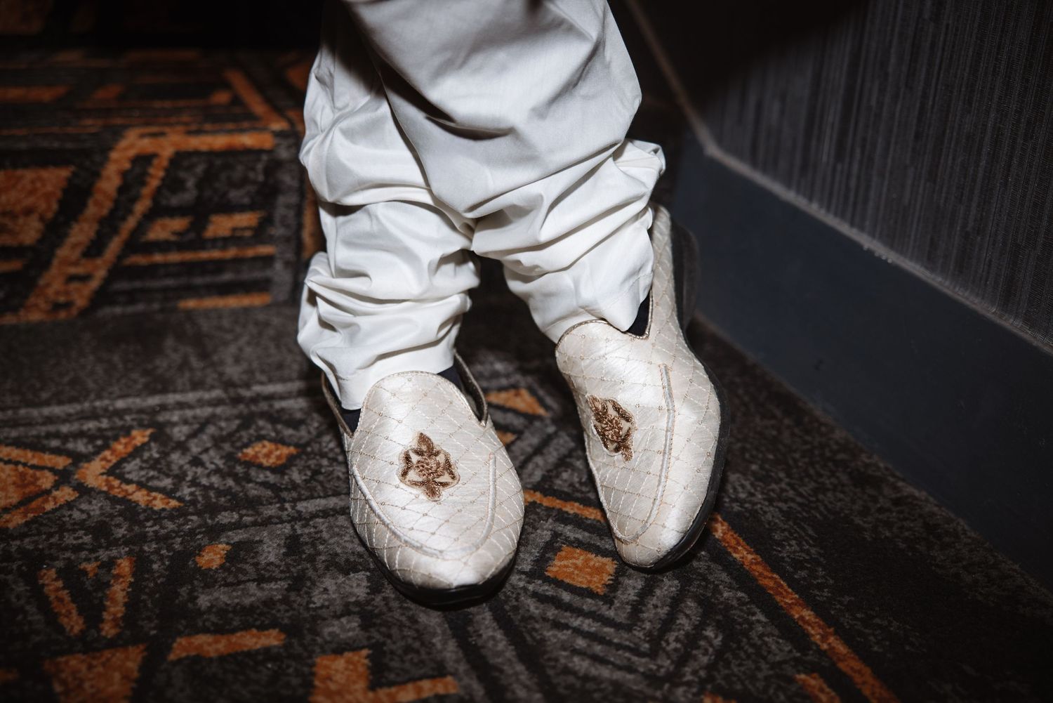 Close-up of groom wearing white embroidered mojari shoes on patterned carpet.
