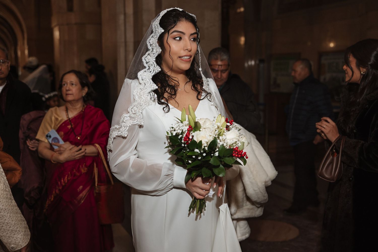 Elegant bride in white long-sleeve dress and lace headpiece carrying red and white floral bouquet.