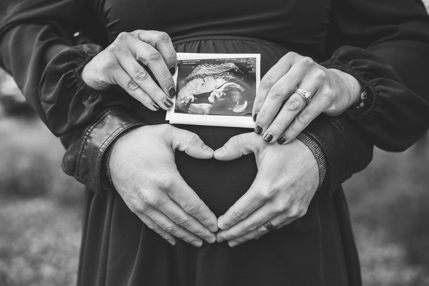 Black and white photo of pregnant couple holding ultrasound image with heart hands on baby bump.