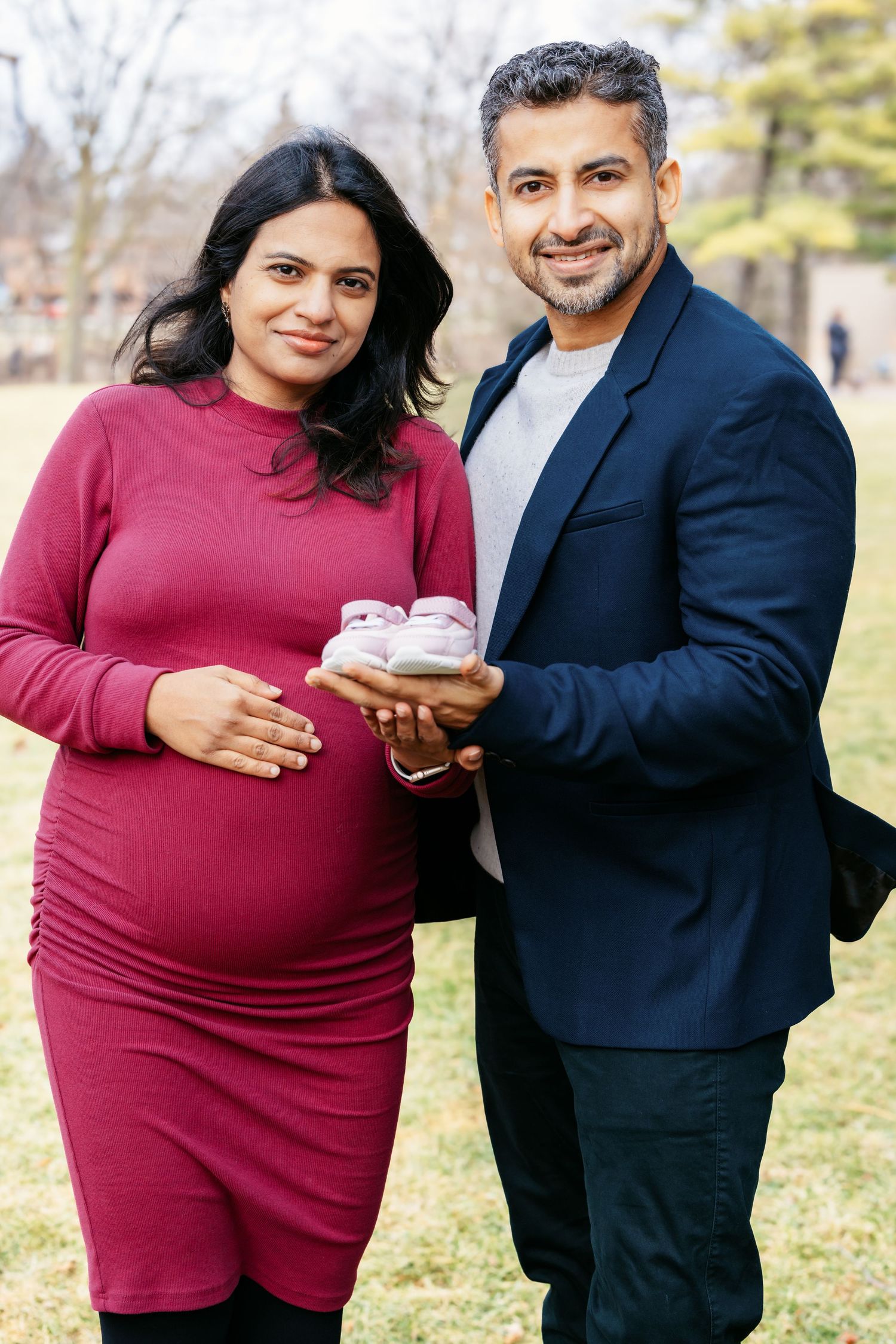 Pregnant woman in red dress and man in navy blazer holding pink baby shoes in a park.
