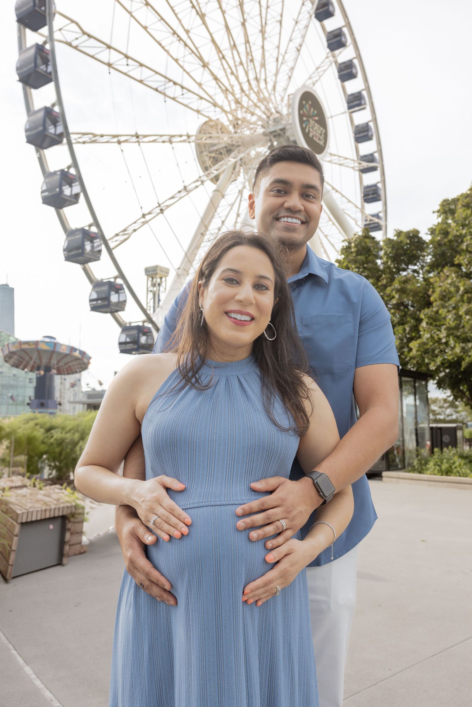 Smiling couple cradling pregnant belly in blue dress with a Ferris wheel in the background outdoors.