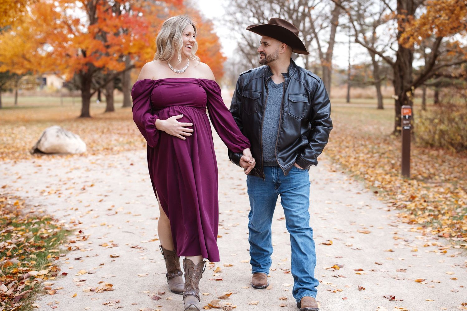 Laughing pregnant woman in off-shoulder dress walks with smiling cowboy partner in fall park.