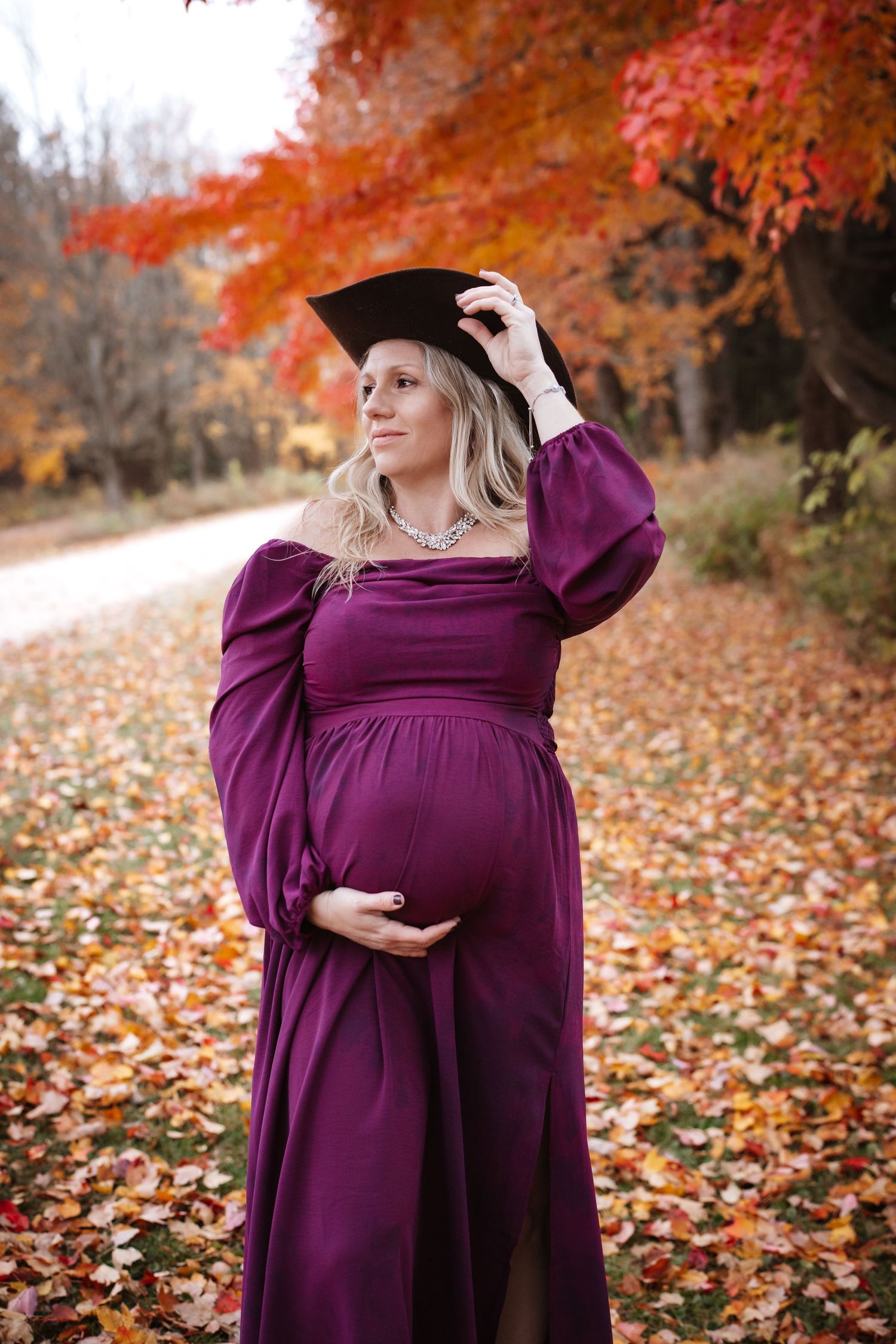 Pregnant blonde woman in purple gown wearing graduation cap in fall foliage.
