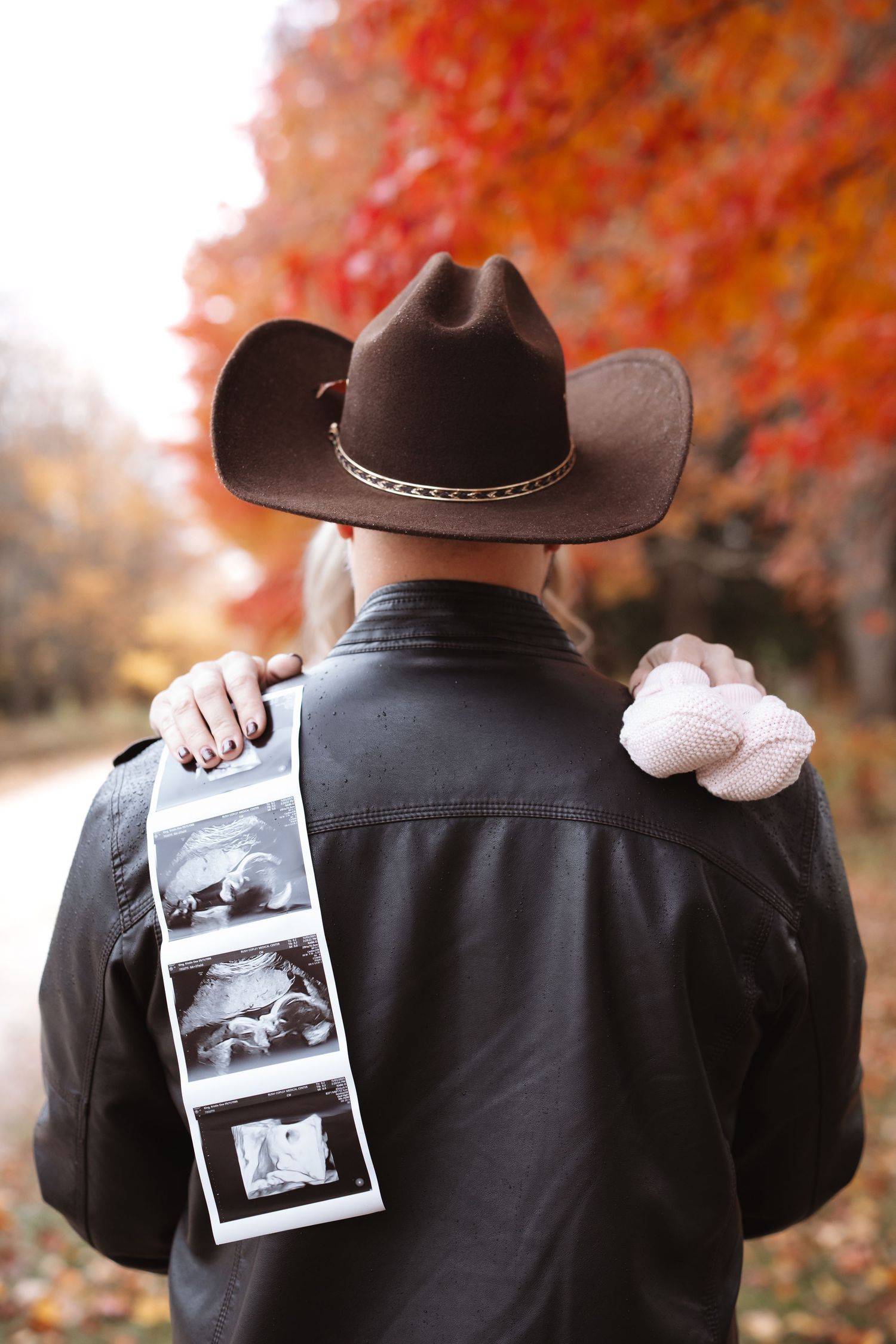 Man in cowboy hat displays ultrasound photos and baby shoes on his back amid fall foliage.