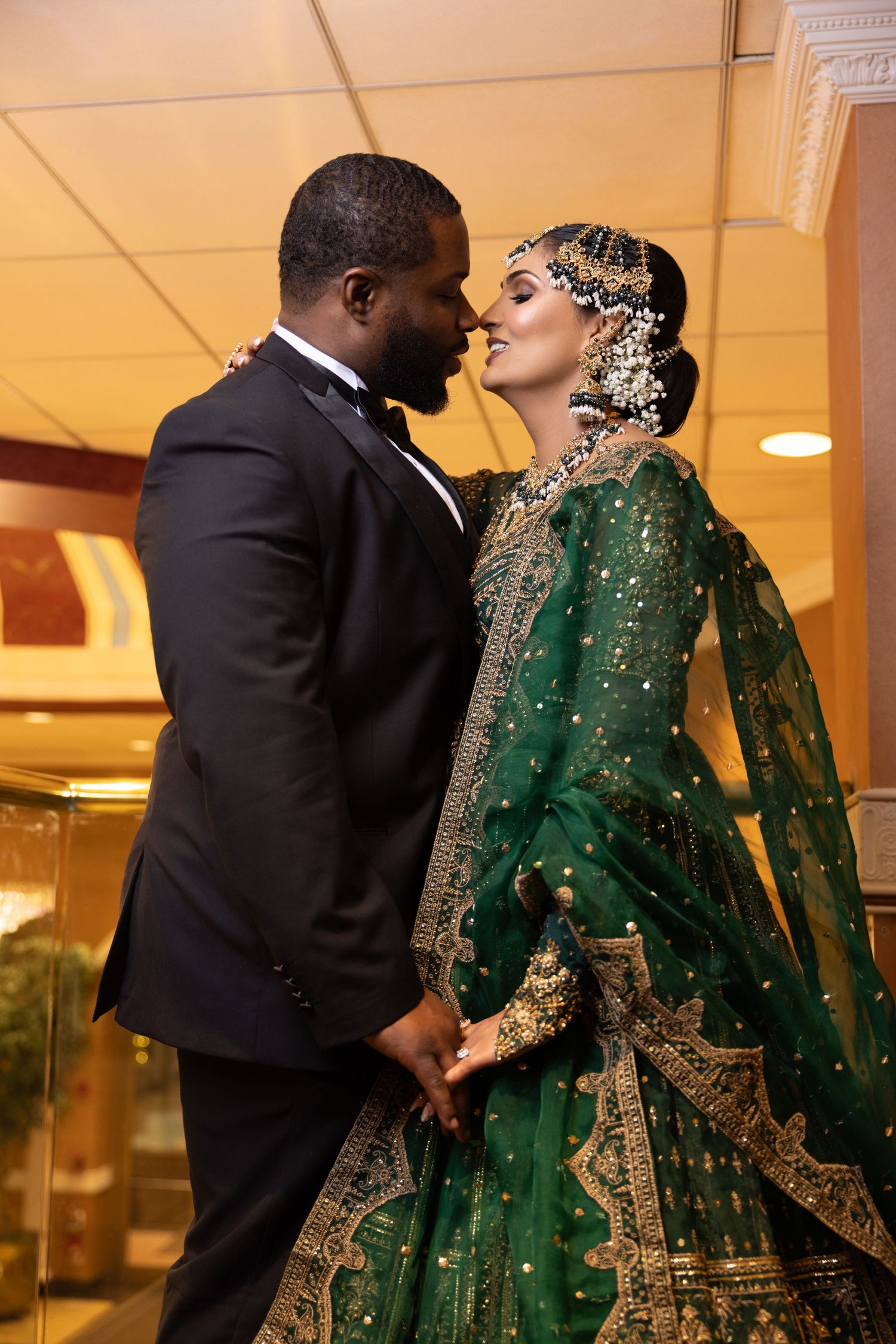 Interracial couple in wedding attire share a romantic moment, bride in green gold-embroidered lehenga.