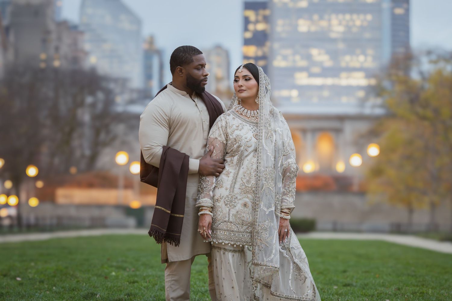 Interracial Muslim couple in traditional South Asian wedding attire posing on grass with city skyline at dusk.