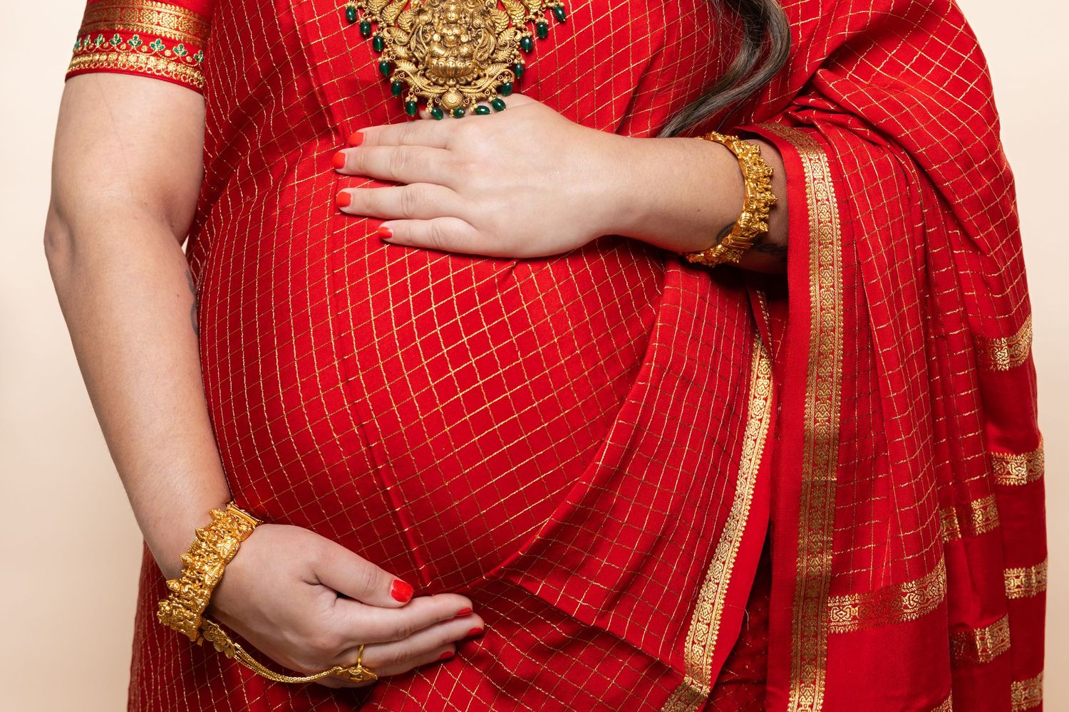 Close-up of pregnant woman in red saree with gold bangles cradling bare baby bump.
