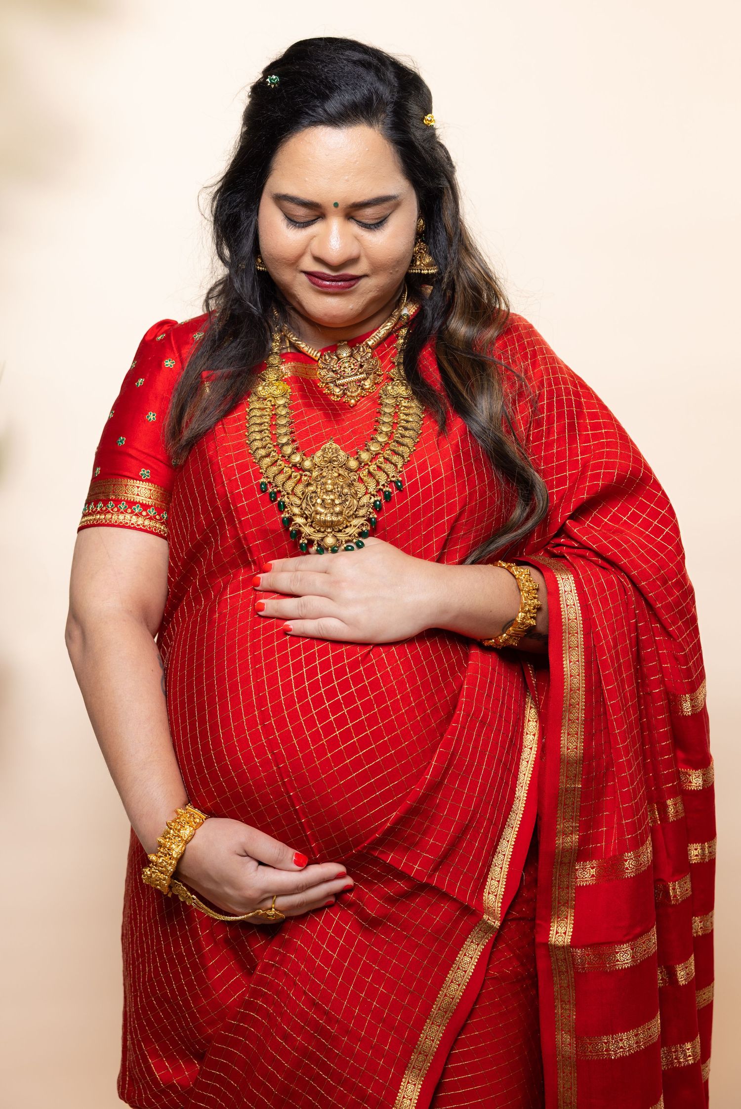 Pregnant Indian woman in red saree with gold jewelry looking down at baby bump.