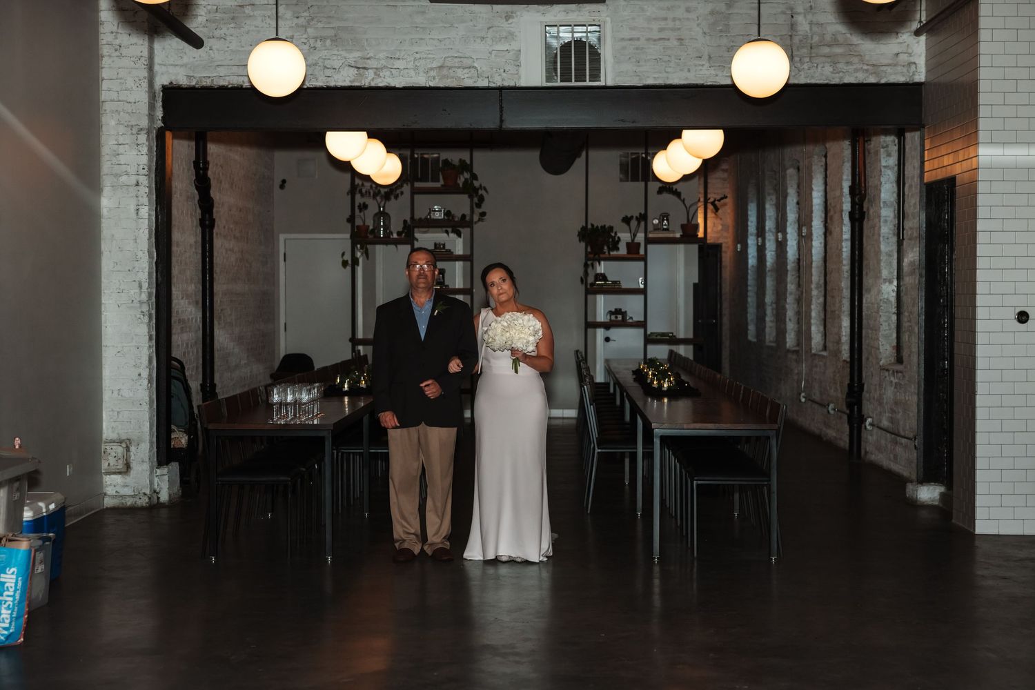 A bride and her escort walk through an elegant industrial wedding venue with brick walls and pendant lights.