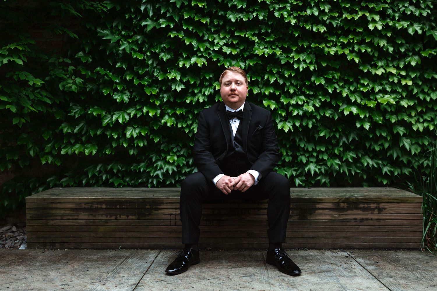 Groom in black tuxedo sits on a wooden bench surrounded by a lush green ivy-covered wall.