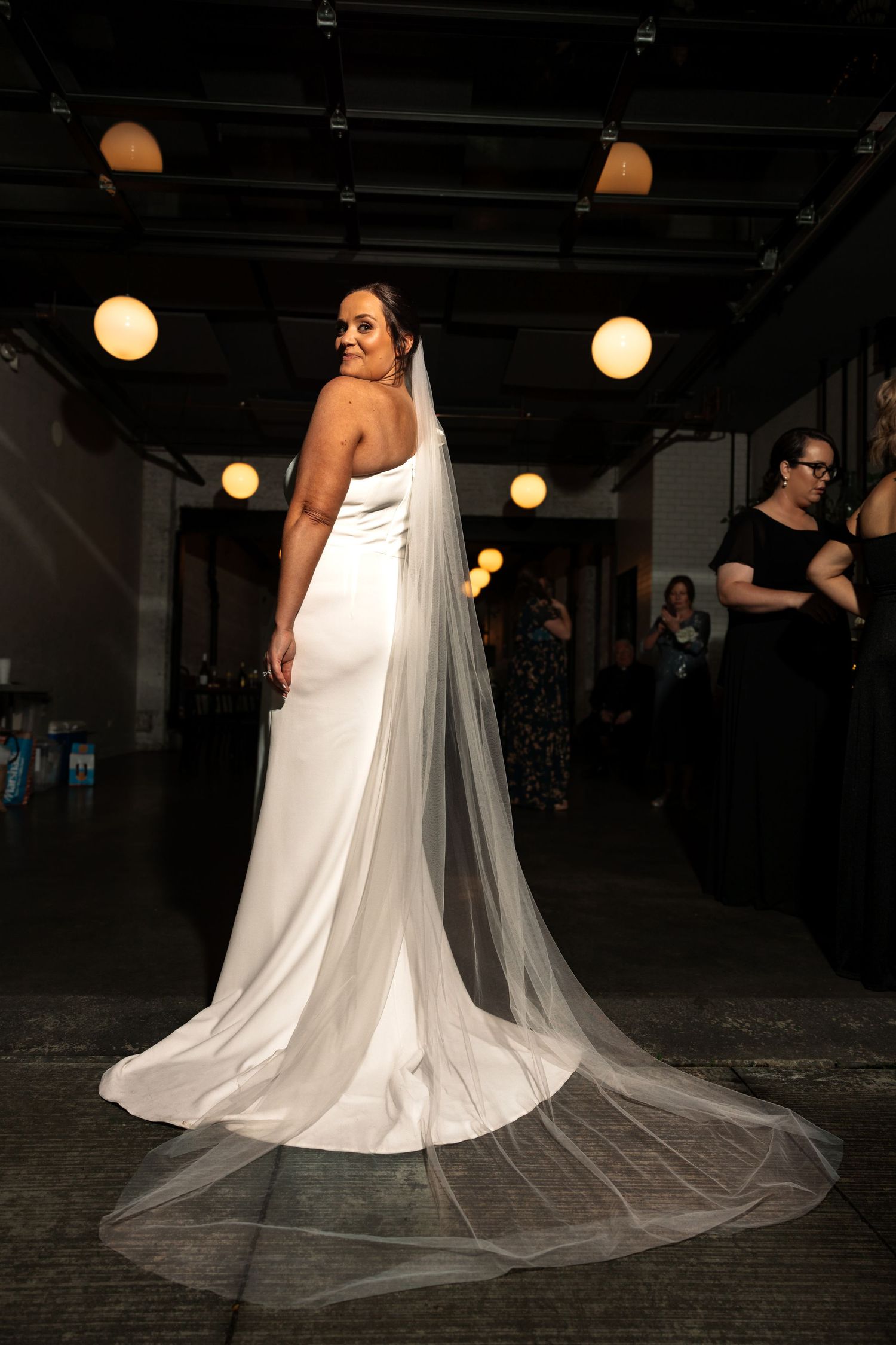 Elegant bride in sleek white gown with dramatic cathedral veil poses from behind in softly lit industrial venue.