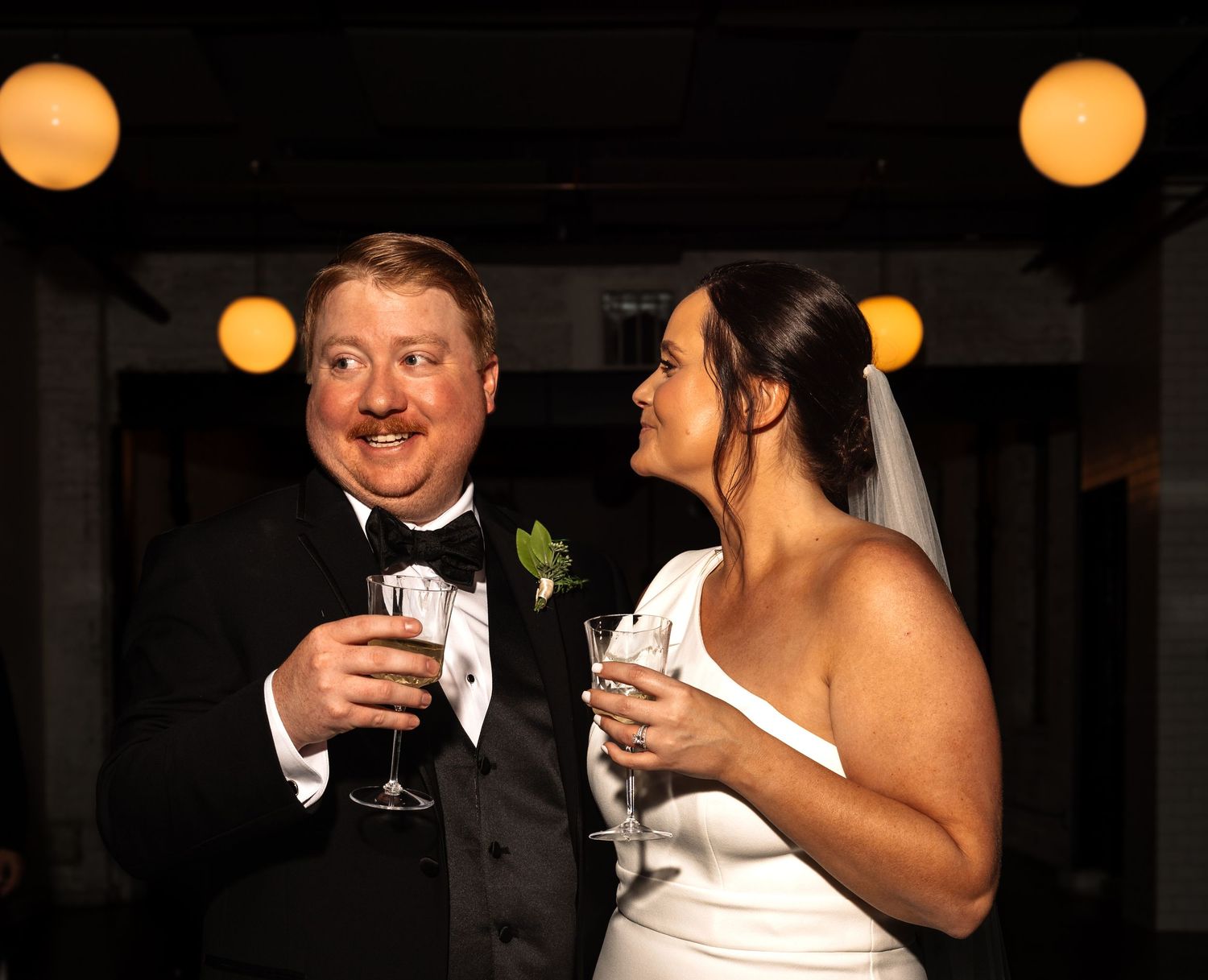 Smiling bride and groom toast with champagne glasses at wedding reception under warm golden lights.