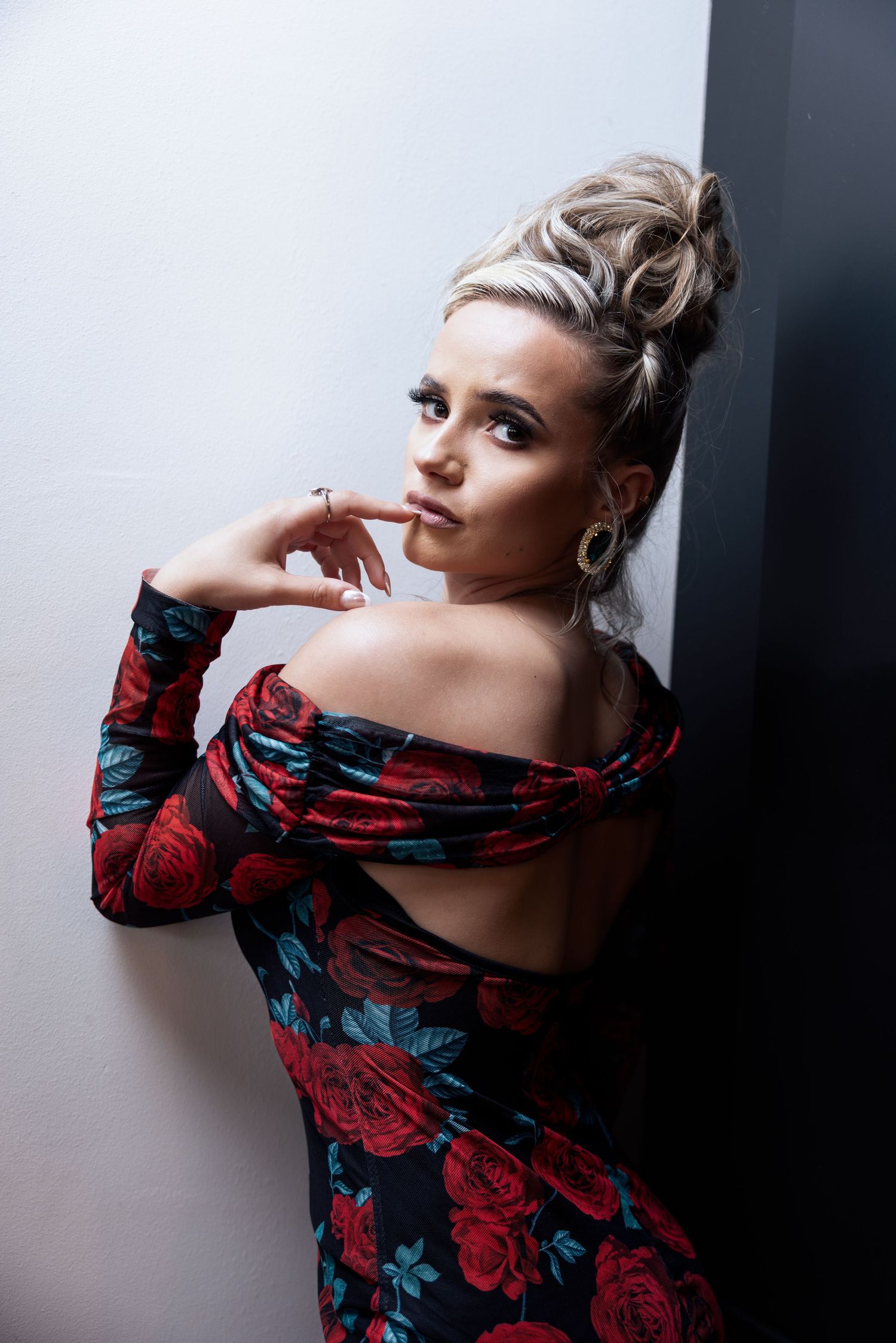 Blonde woman in off-shoulder red floral dress with updo posing against dark wall.