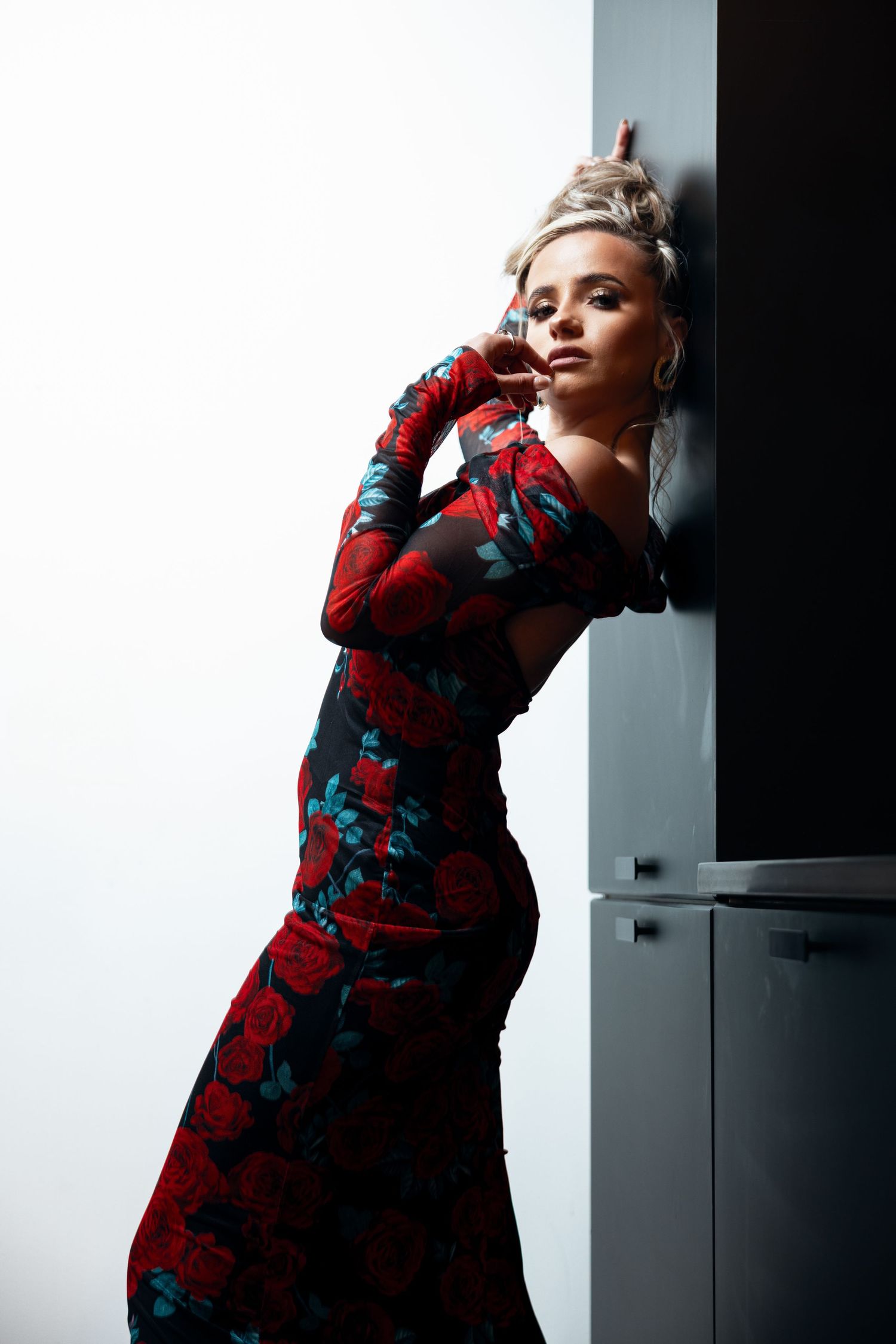 Blonde woman in floral red rose print form-fitting gown leans against dark wall in dramatic pose.