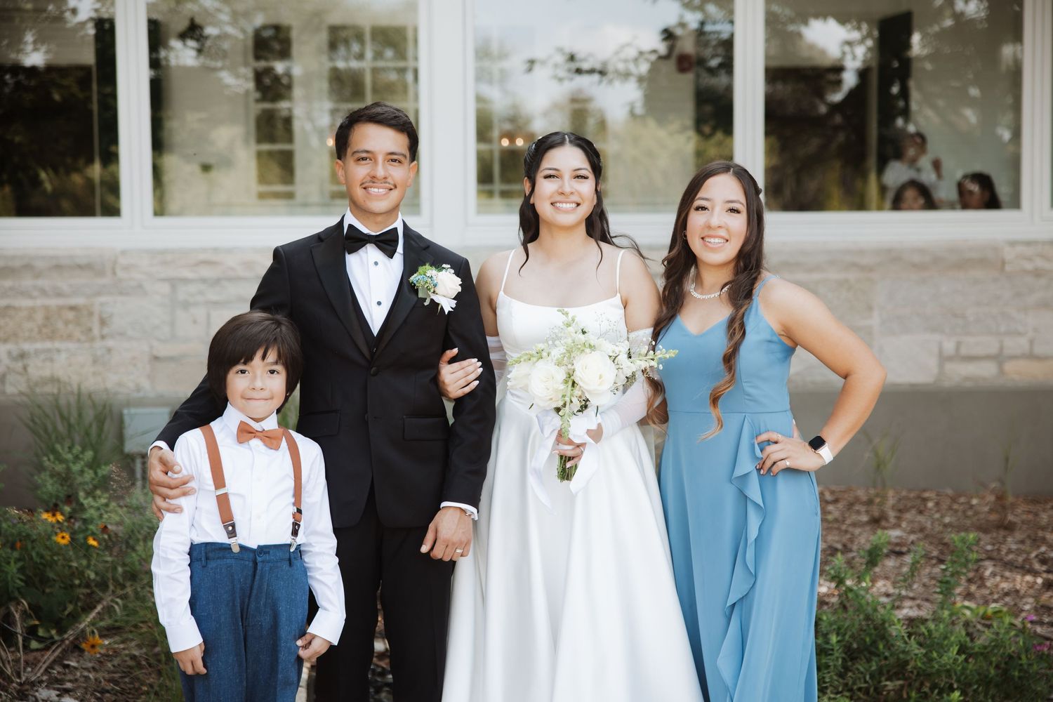 Wedding couple smiles with ring bearer in suspenders and bridesmaid in dusty blue gown outdoors.