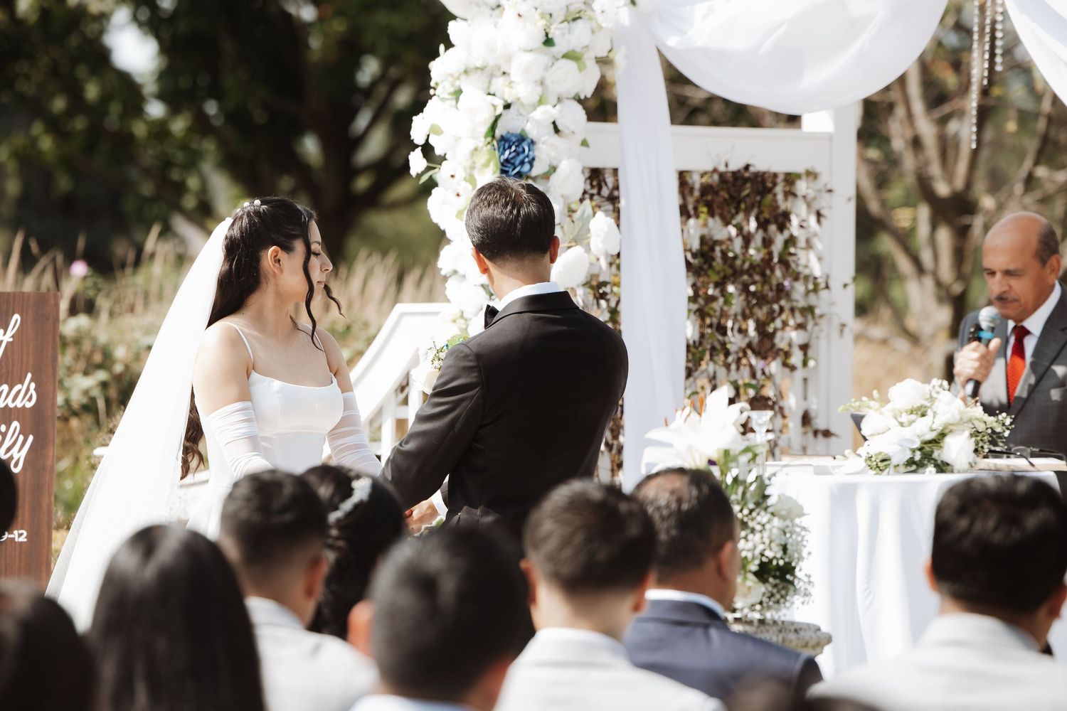 Bride and groom at altar during outdoor wedding ceremony with white floral arch backdrop.