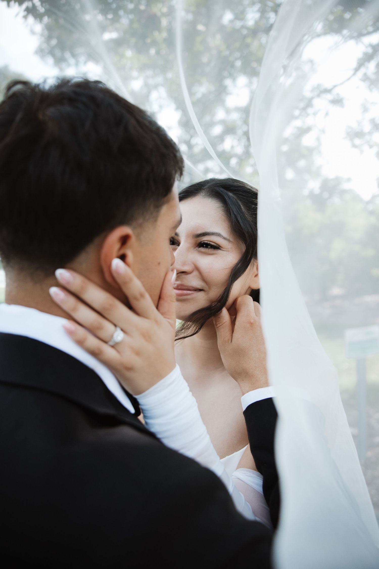 Smiling bride gazes at groom under flowing white veil, gently holding his face with diamond ring visible.