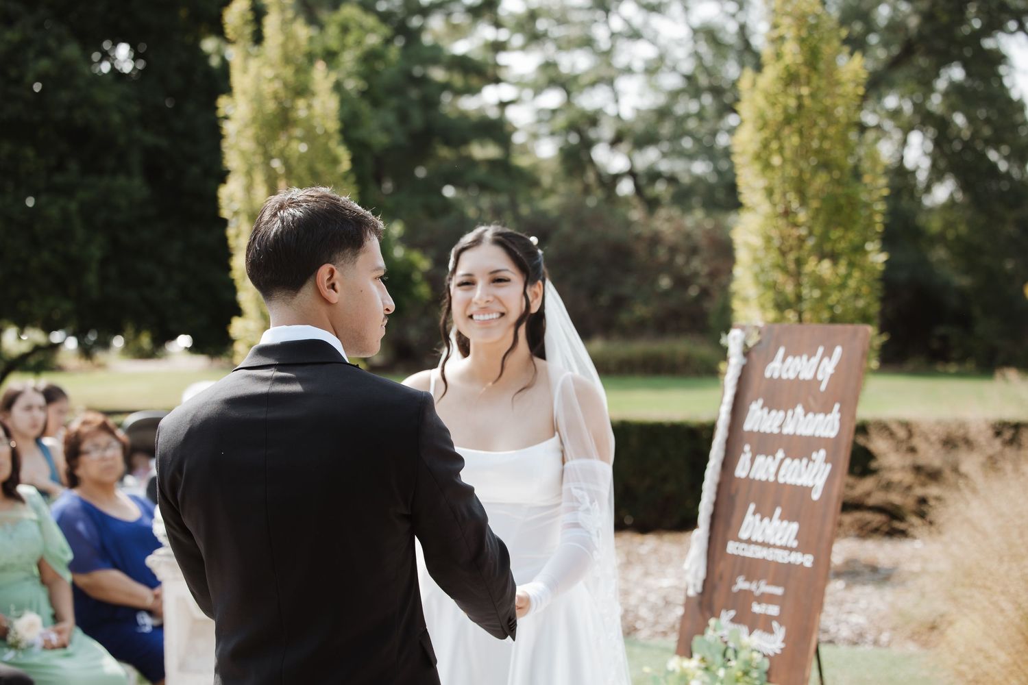 Bride and groom exchange vows at outdoor wedding ceremony with wooden sign and seated guests.