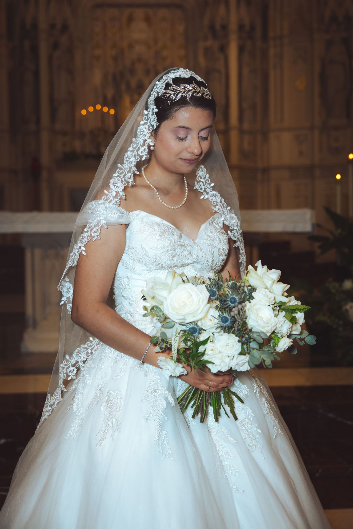 Bride in white lace gown holding bouquet of white roses in church.