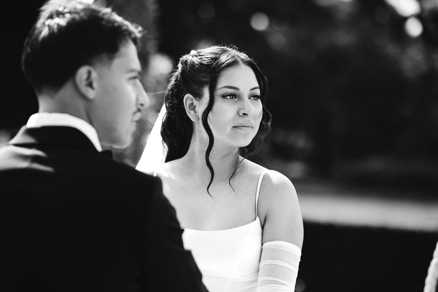 A black and white portrait of a bride gazing at the groom during their outdoor wedding ceremony.