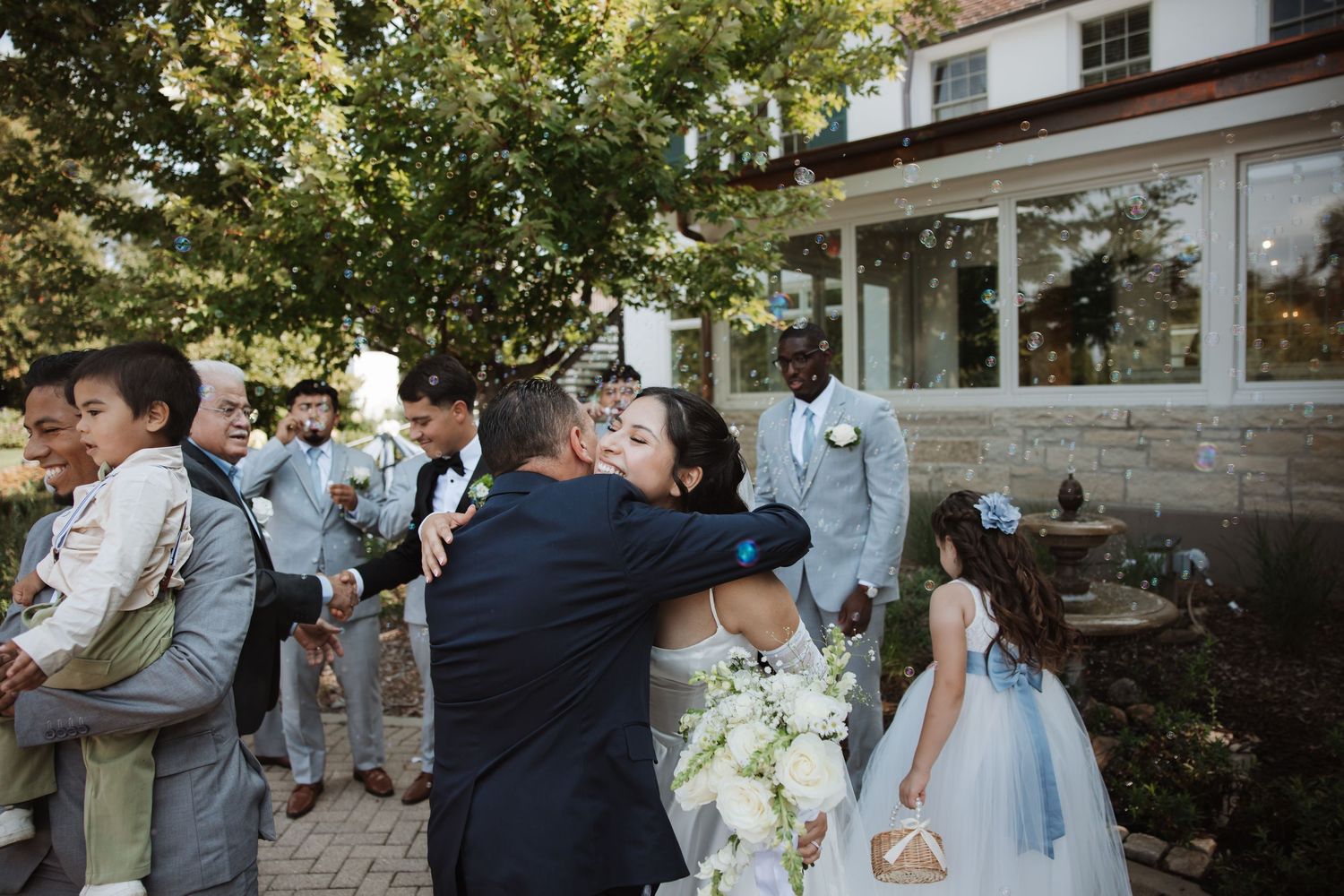 Bride and groom greeting family members with hugs outside a venue surrounded by wedding party members.