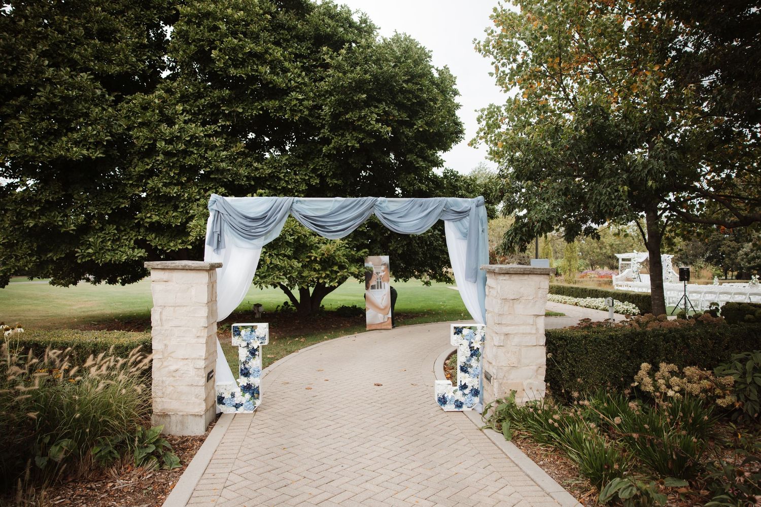 Stone pillar wedding entrance arch with blue draping and floral decorations along a brick garden pathway.