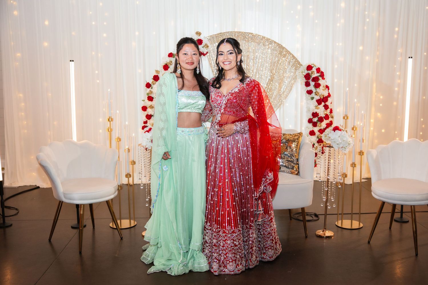 Two women in elegant Indian traditional outfits posing together at a beautifully decorated wedding celebration venue.