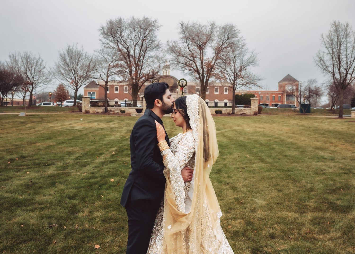 South Asian bride and groom share romantic kiss outdoors in winter garden wearing traditional wedding attire.