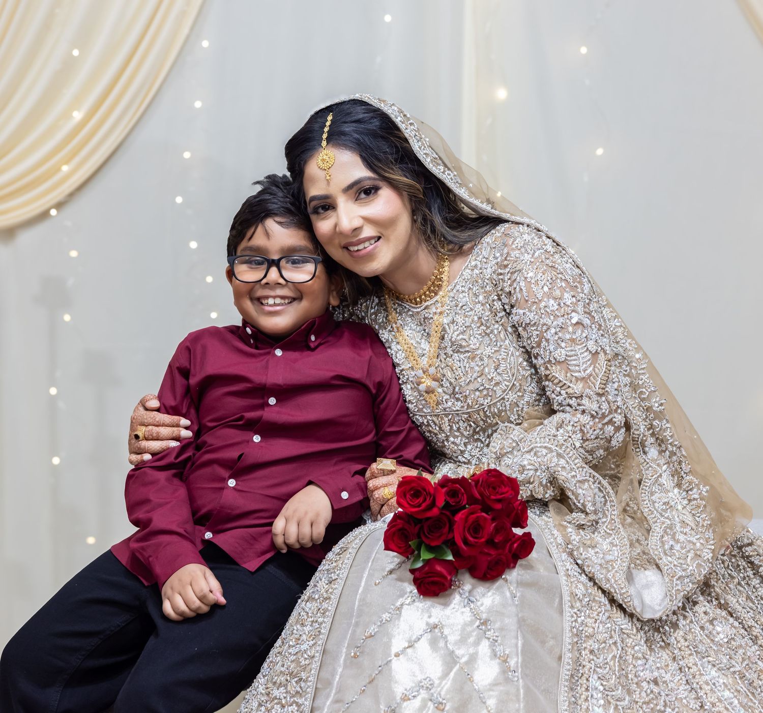 Bride in gold bridal lehenga smiles with young boy in maroon shirt holding red roses at wedding backdrop.