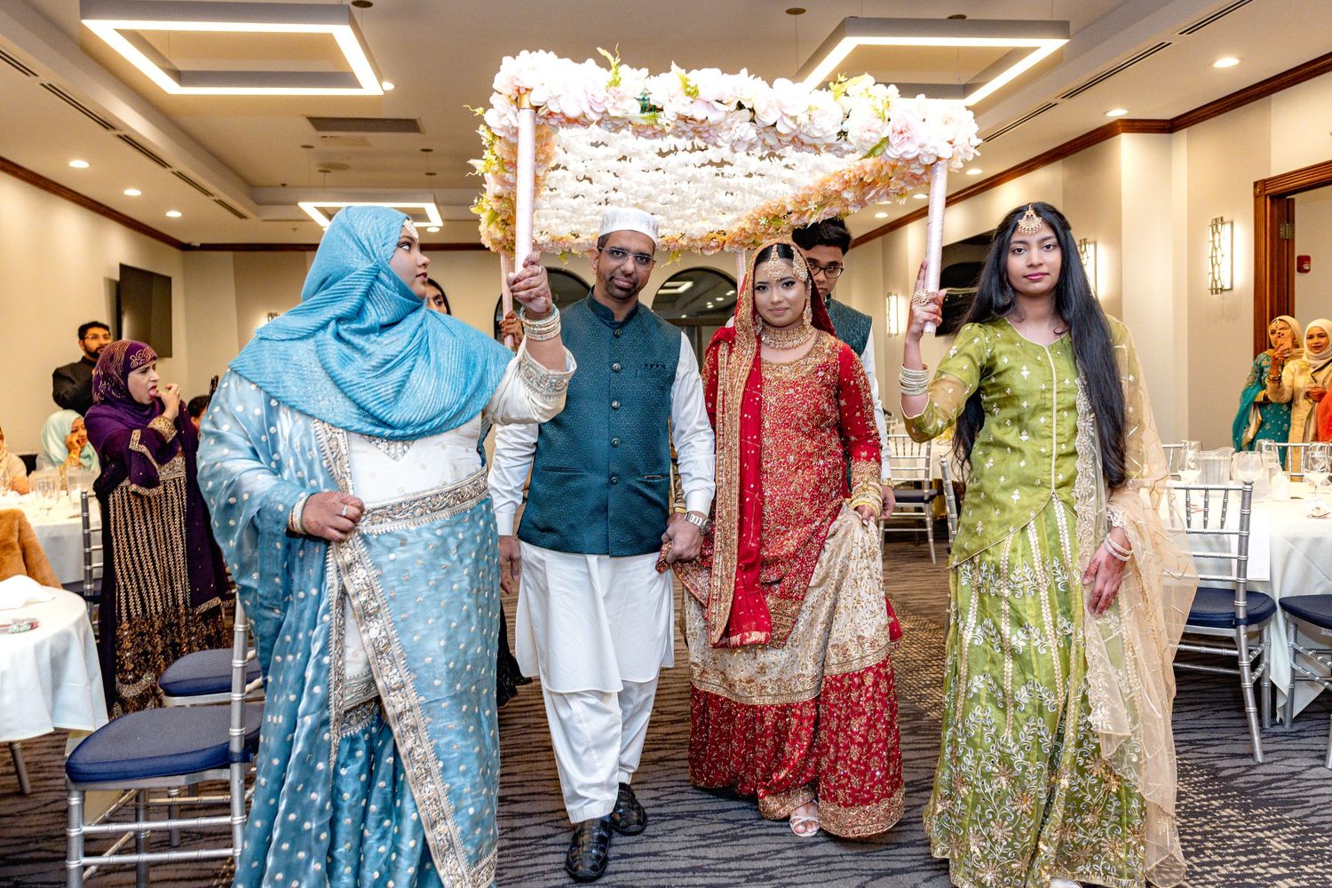 South Asian bride in red bridal lehenga escorted under floral canopy by family at wedding reception.