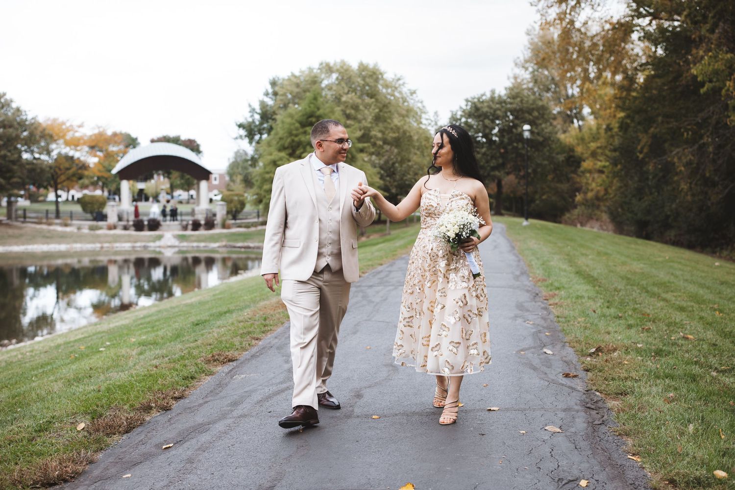 Couple holding hands walking on a paved path near a pond and gazebo in a park on their wedding day.