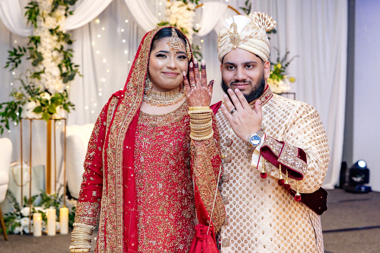 South Asian bride and groom in traditional red and gold wedding attire pose together showing wedding rings.