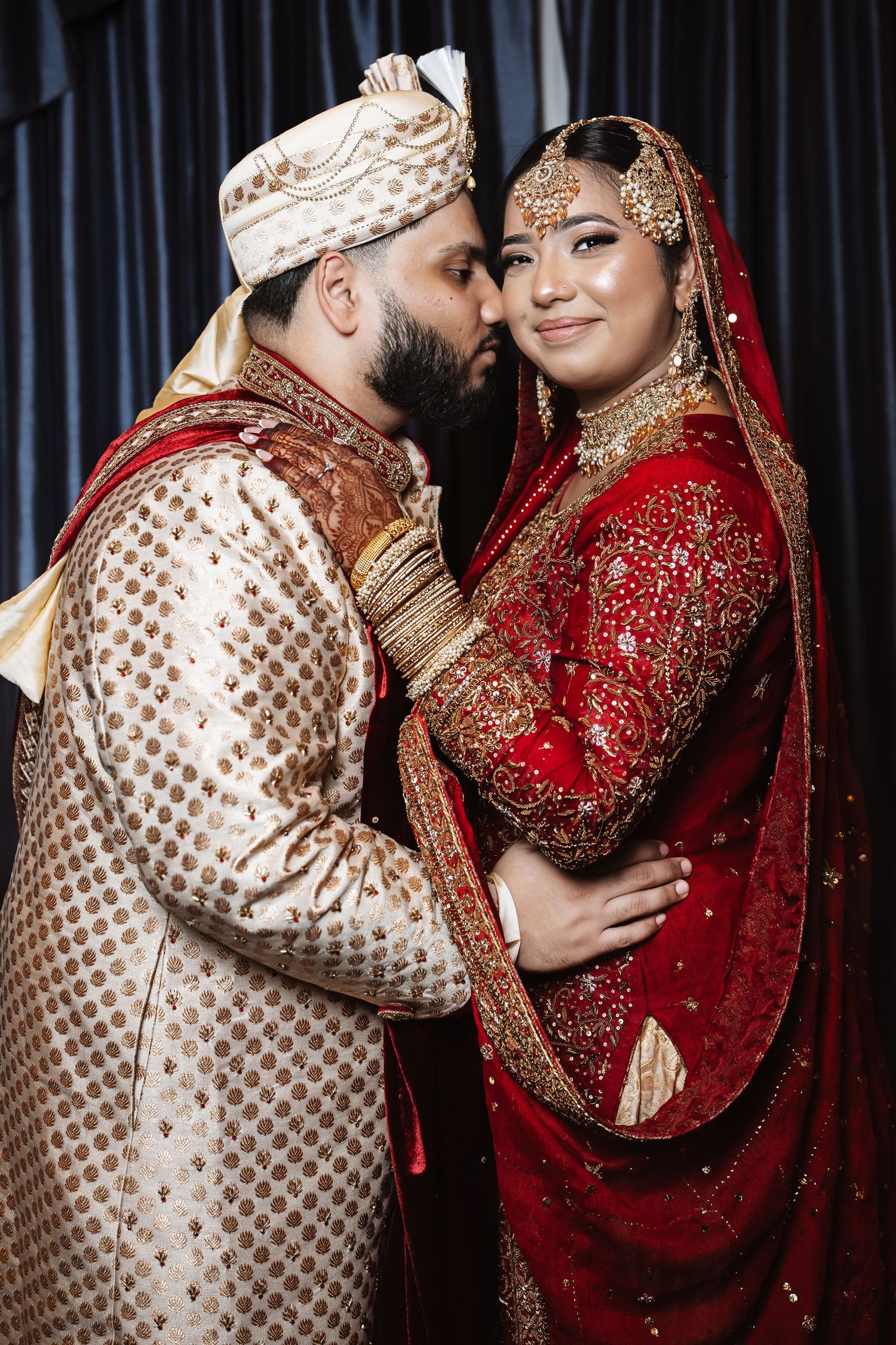 South Asian groom in gold sherwani kisses bride in red bridal lehenga with gold jewelry at wedding.