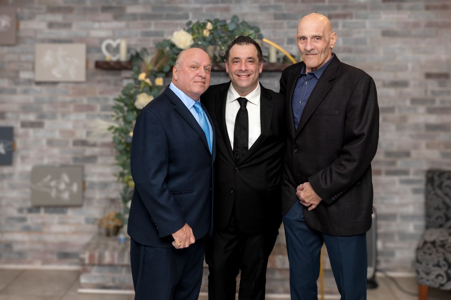 Three men in suits smiling together at a formal event with stone wall backdrop.