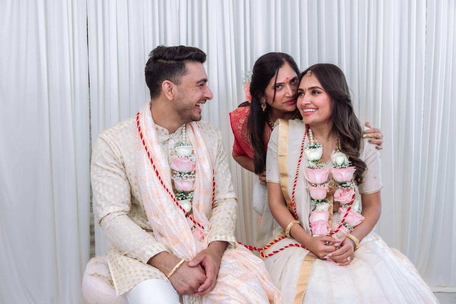 Indian newlyweds in garlands share joyful moment with woman in red saree between them.