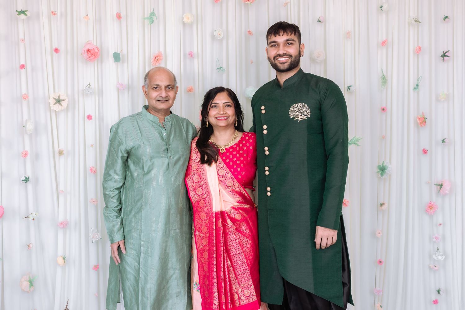 Family of three in elegant Indian traditional wear smile together at festive celebration with floral backdrop.