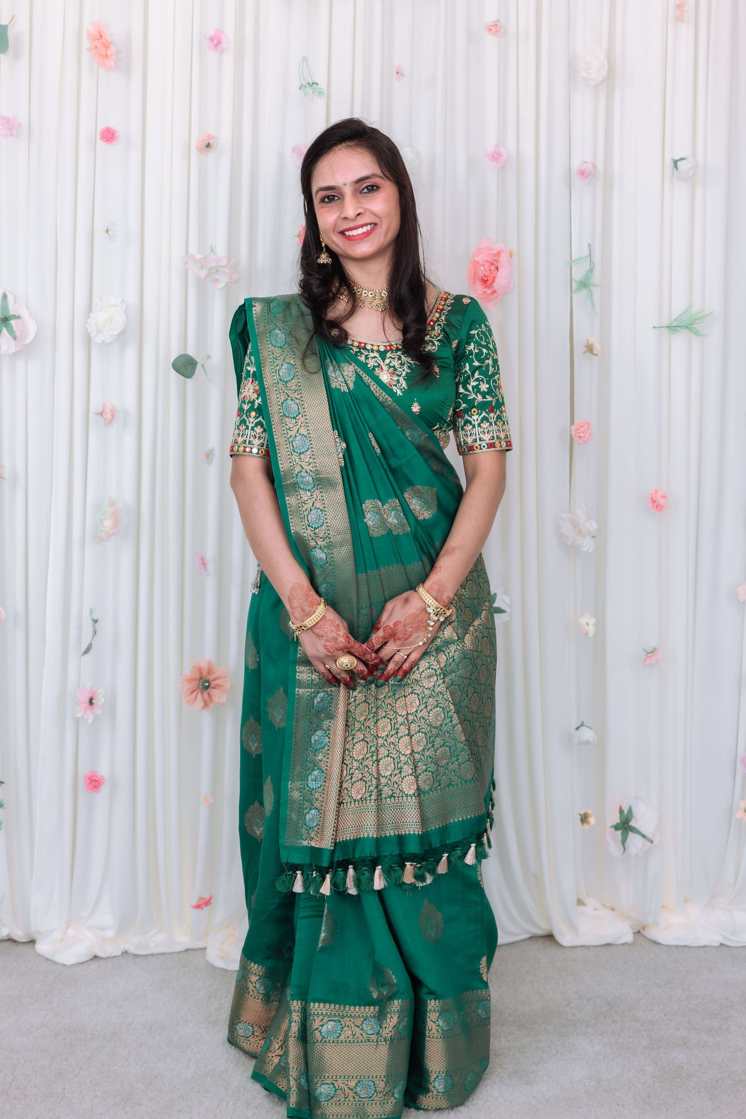 Woman smiling in elegant green Banarasi silk saree with gold zari work, posing in front of white floral backdrop.