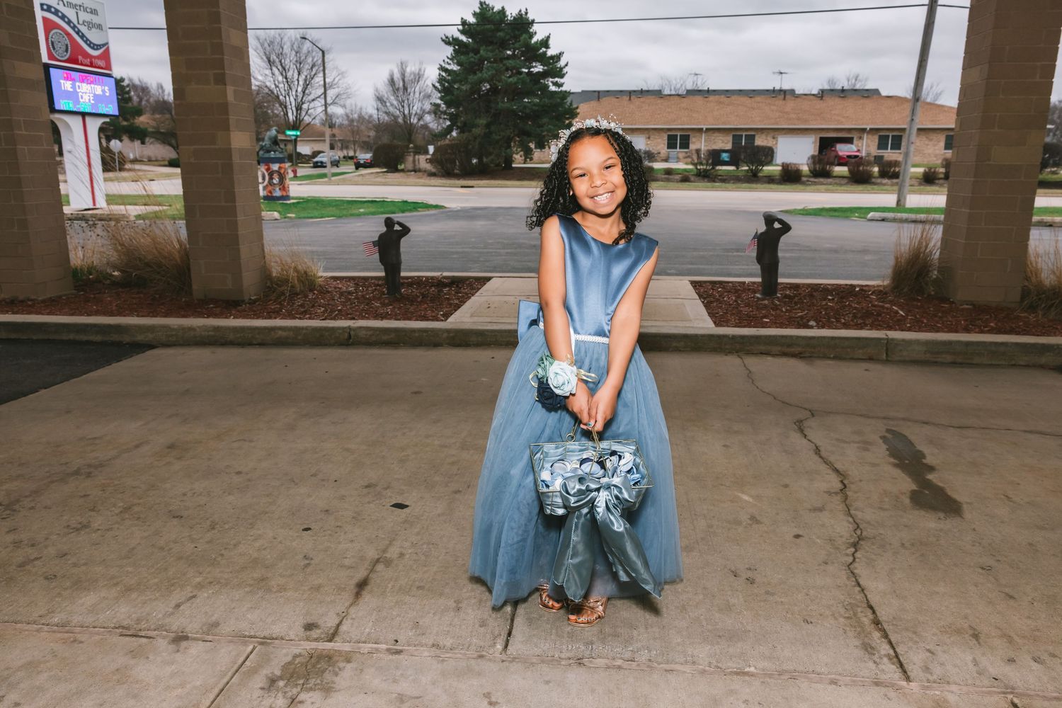 Smiling flower girl in blue gown holding bouquet standing outside wedding venue.