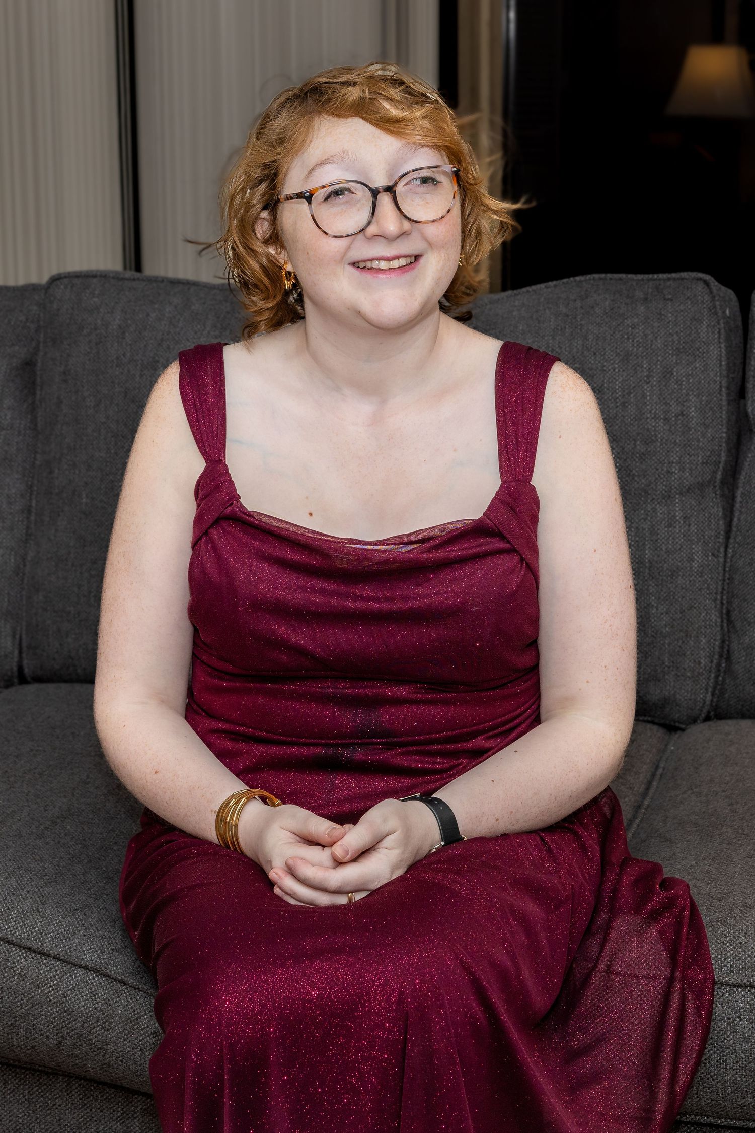 Smiling redhead woman wearing glasses and burgundy sparkle gown seated on gray sofa.